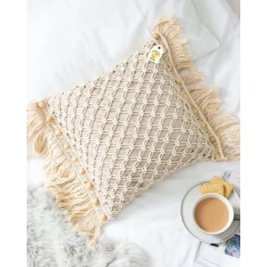 Cream cushion cover with an open diamond pattern, wooden-tone beads and fringe on white bedding.